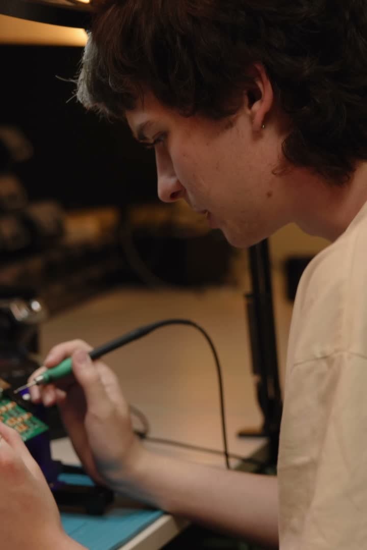 Bálint soldering a circuit board on the bench, test equipment blurred behind it.