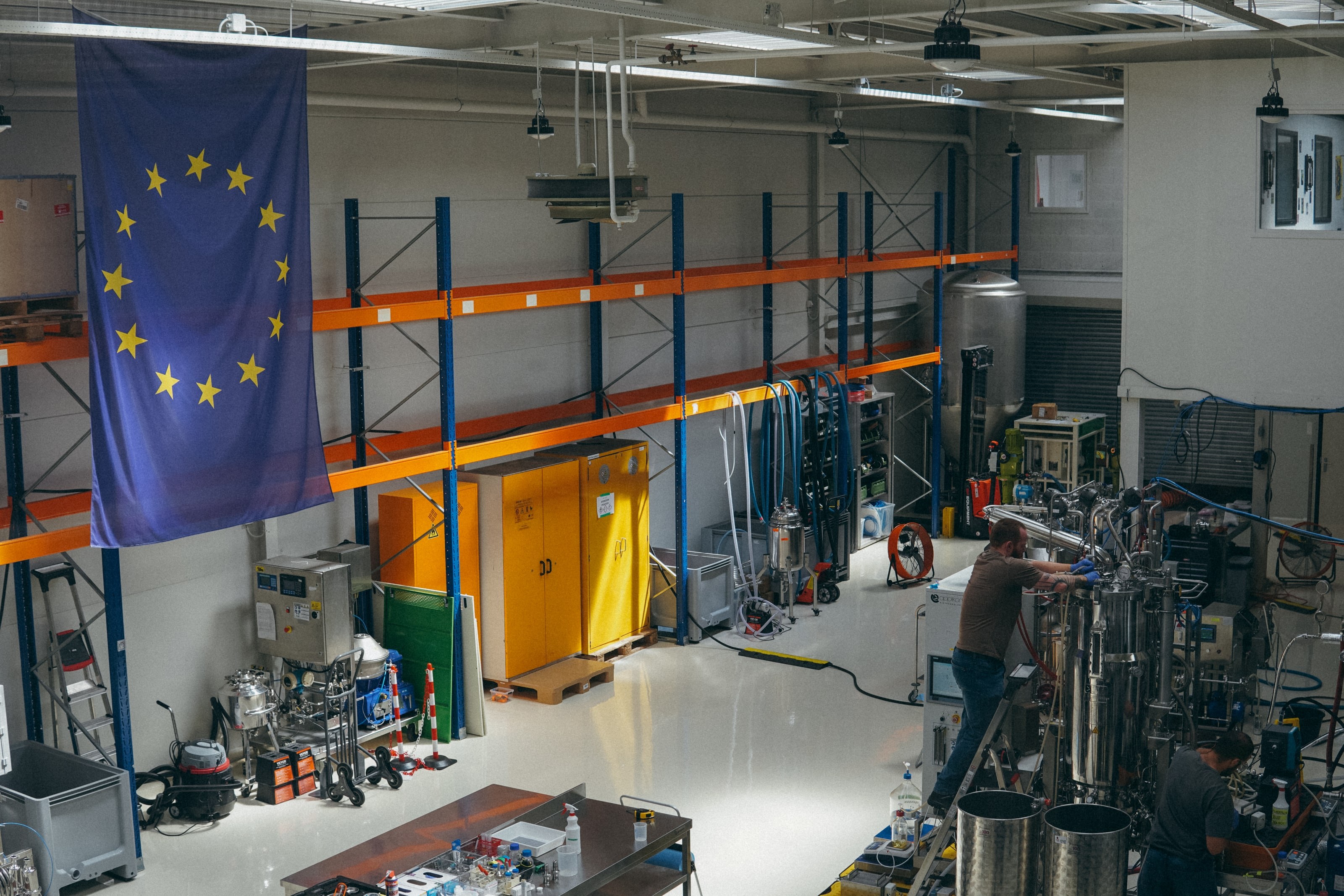 The pilot brewery on the Valschermkade: an EU flag over the hall, one person working at the fermentation skid.