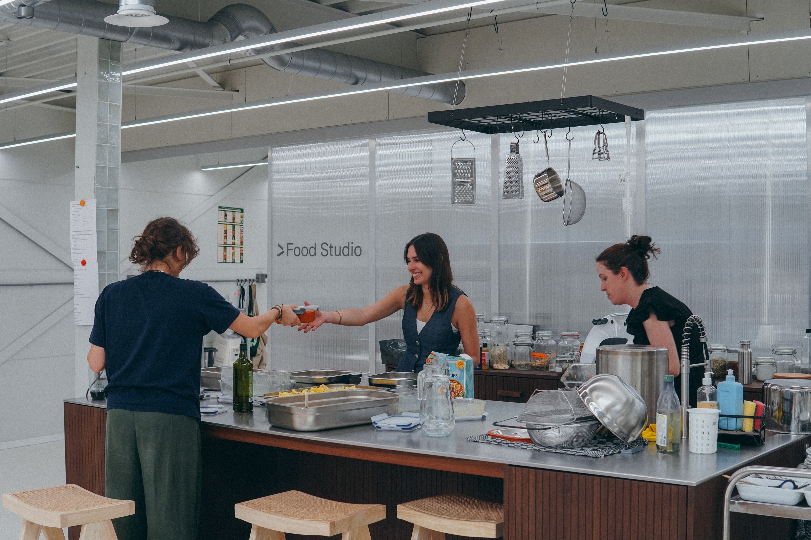 Three colleagues cooking lunch together in the Food Studio, one handing a tray across the bench.