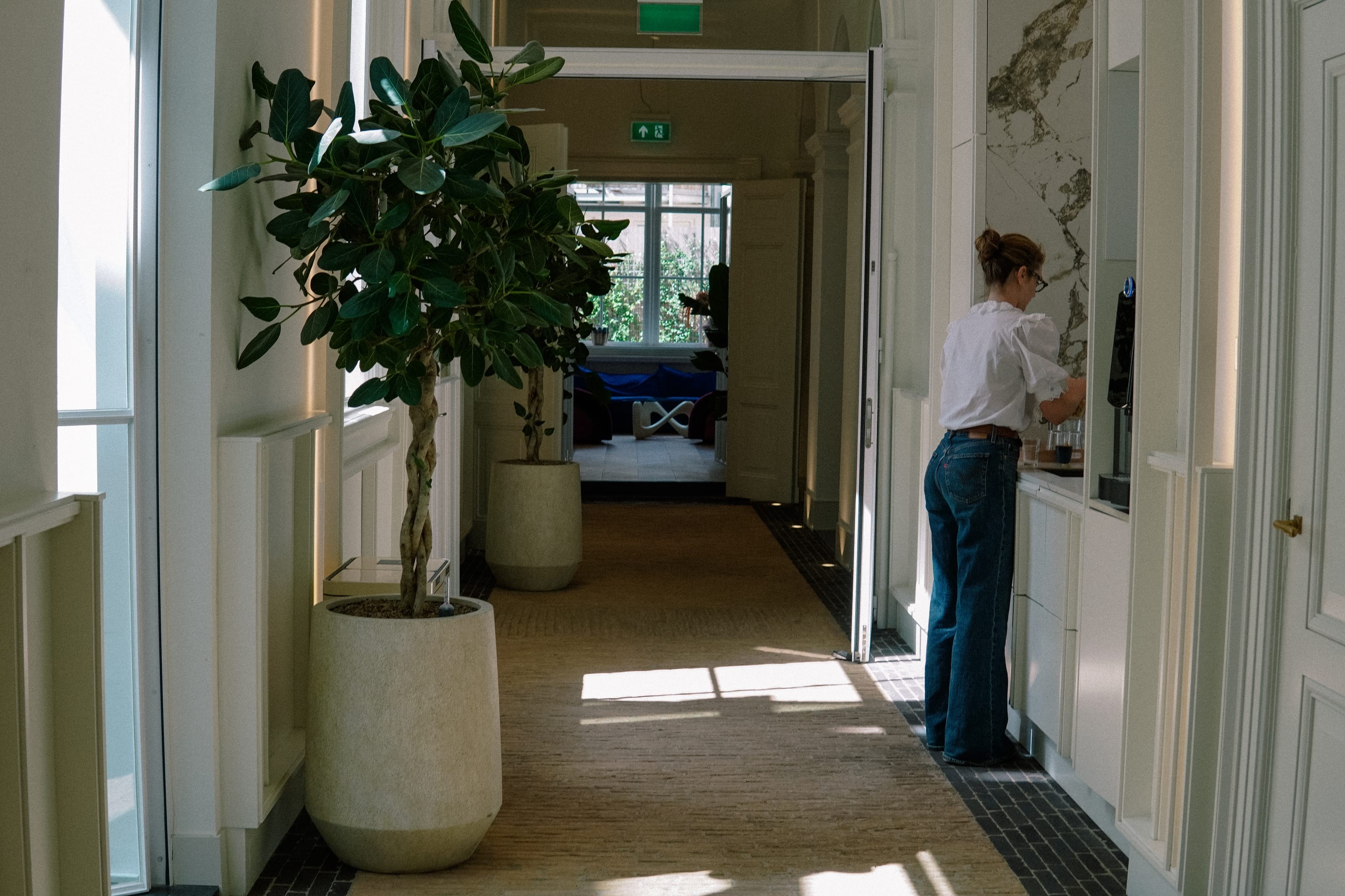 A corridor with a large ficus in a stone pot, and a woman at a coffee machine on a marble counter at the far end.