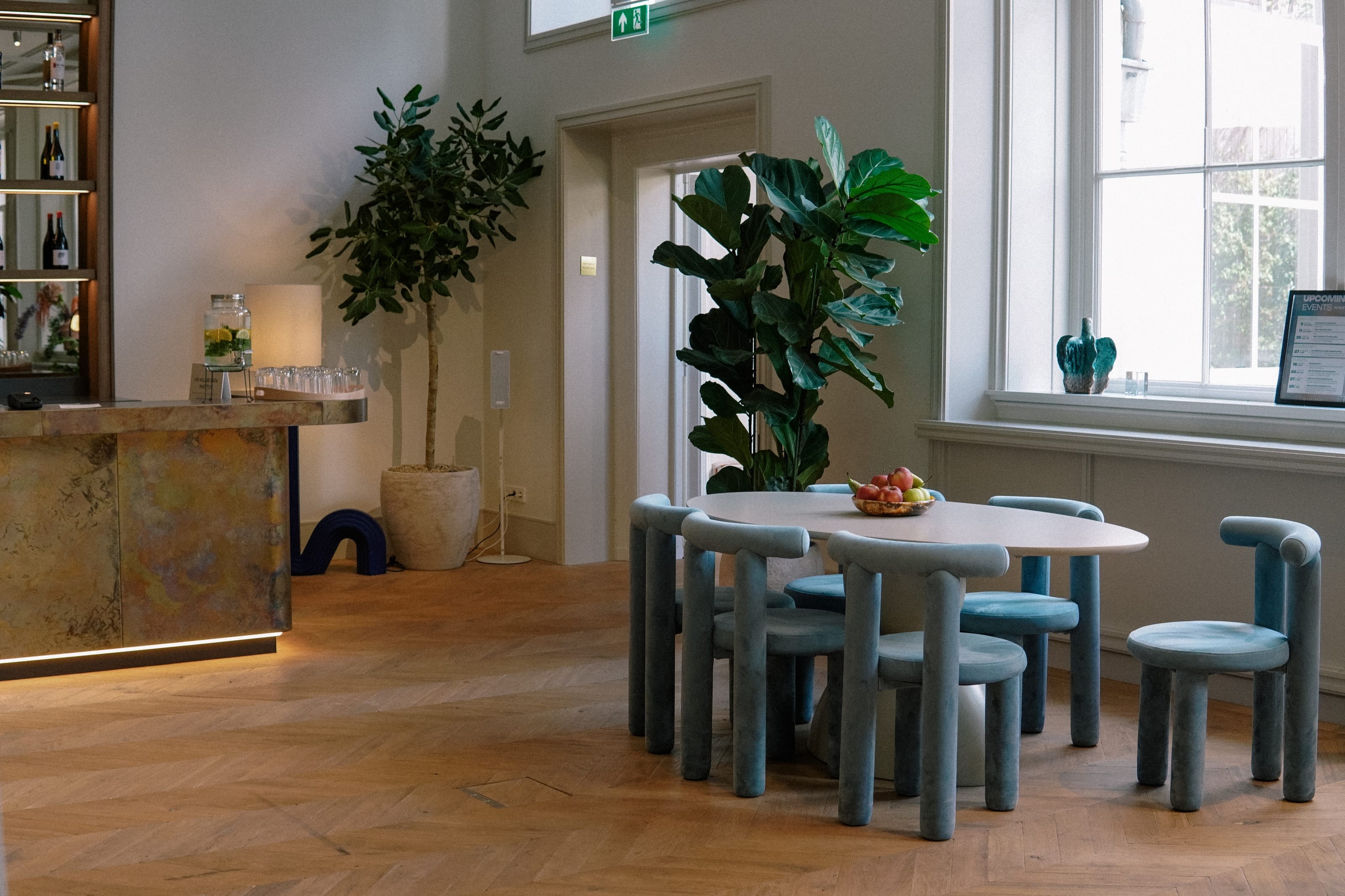 A round table with blue velvet chairs and a bowl of fruit, a bar counter and plants beside it.