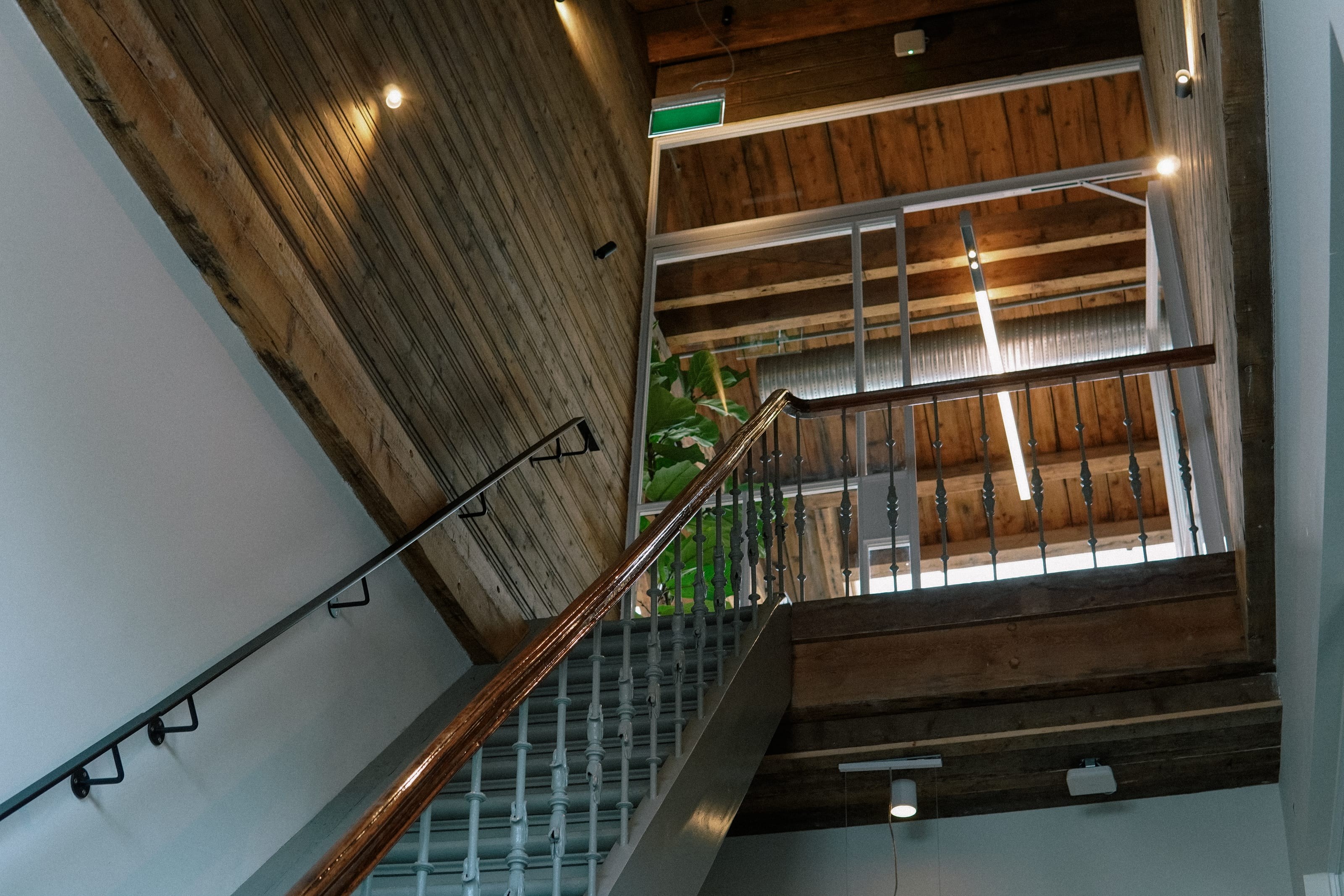 Looking up a stairwell with a wooden banister, under a wooden ceiling with exposed beams.